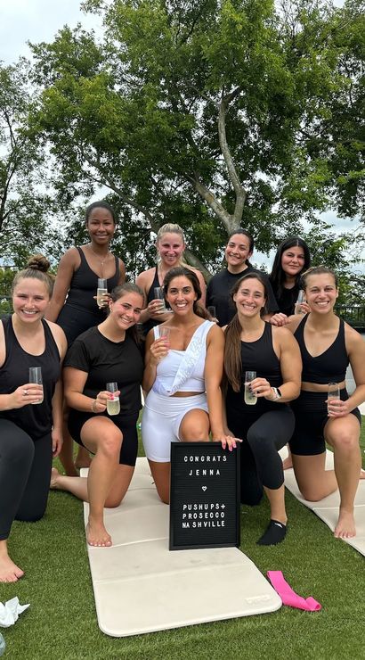 Rooftop fitness celebration in Nashville: nine women in activewear pose on outdoor yoga mats, smiling and toasting with prosecco beside a congratulatory letter board, with trees in the background.