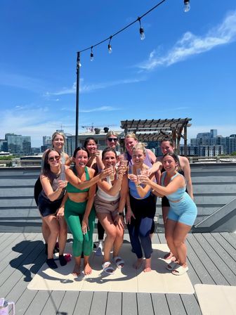 Group of women in colorful activewear toasting drinks on a sunny urban rooftop over yoga mats, with city skyline, string lights and a pergola in the background.
