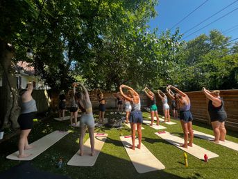 Sunlit backyard yoga class – a group stretching on mats under a large shady tree on green turf in a suburban yard.