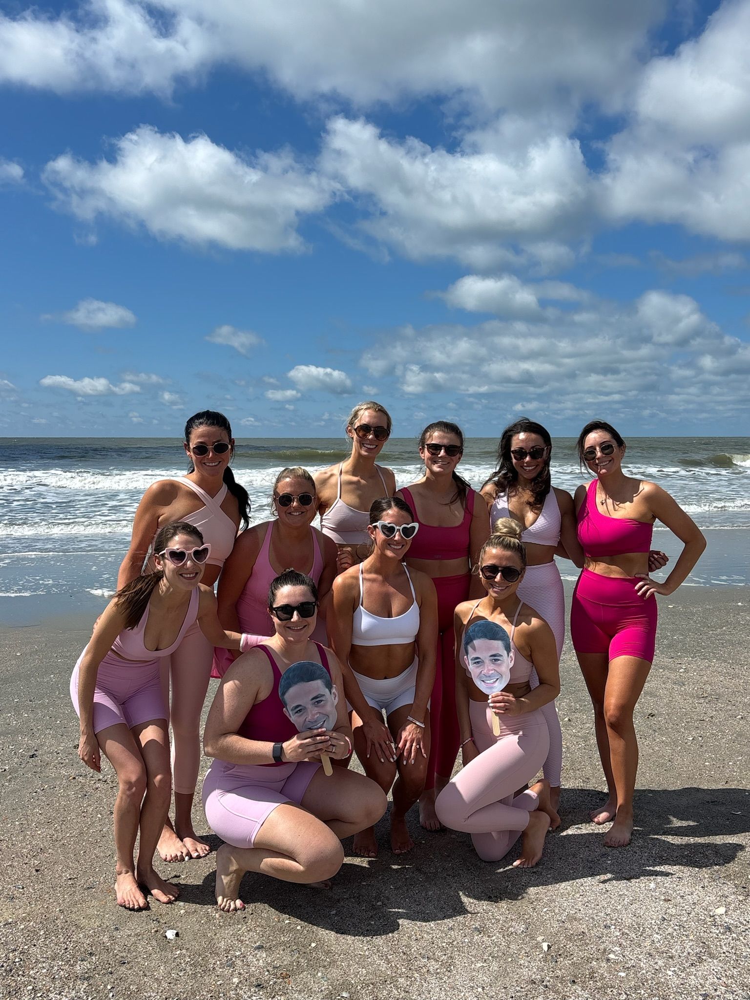 Group of women in pink activewear posing on a sunny ocean beach with blue sky and white clouds, many wearing sunglasses and two holding cardboard face cutouts.