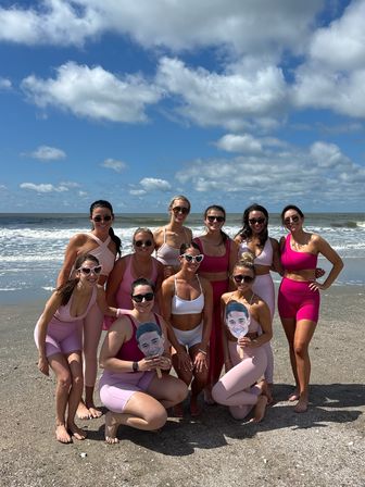 Group of women in pink activewear posing on a sunny ocean beach with blue sky and white clouds, many wearing sunglasses and two holding cardboard face cutouts.