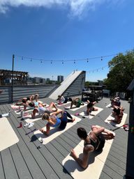Sunny urban rooftop yoga class with participants on exercise mats stretching in side-plank and seated poses, string lights overhead and a downtown skyline in the background