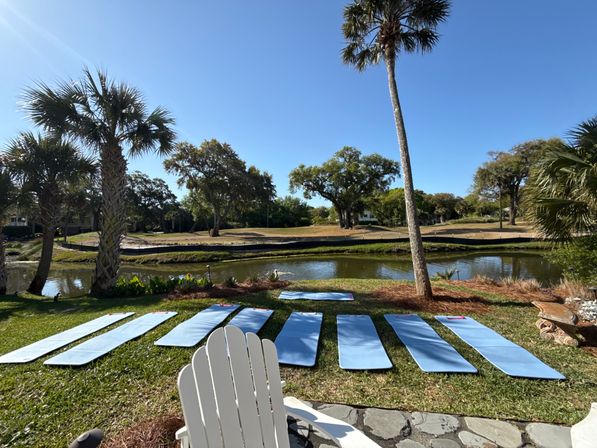 Blue yoga mats laid out on a grassy lawn by a calm pond, framed by palm trees and a bright blue sky for a relaxed outdoor lakeside yoga setup in a tropical coastal park.