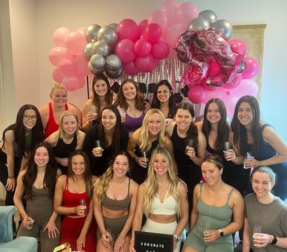 Group of women in activewear posing indoors under a pink-and-silver balloon arch and sequin backdrop, holding champagne flutes and a congratulatory sign with gift bags — celebratory fitness gathering in Nashville.