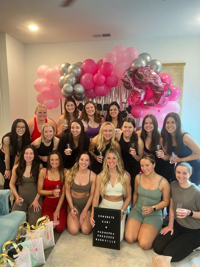 Group of women in activewear posing indoors under a pink-and-silver balloon arch and sequin backdrop, holding champagne flutes and a congratulatory sign with gift bags — celebratory fitness gathering in Nashville.