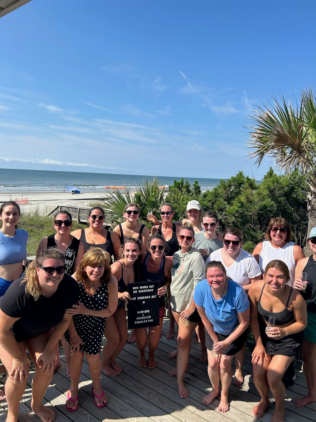 Smiling group of women on a sunny oceanfront wooden deck with palm trees and sandy beach in the background, one holding a small letter board — coastal Charleston beach vibe.