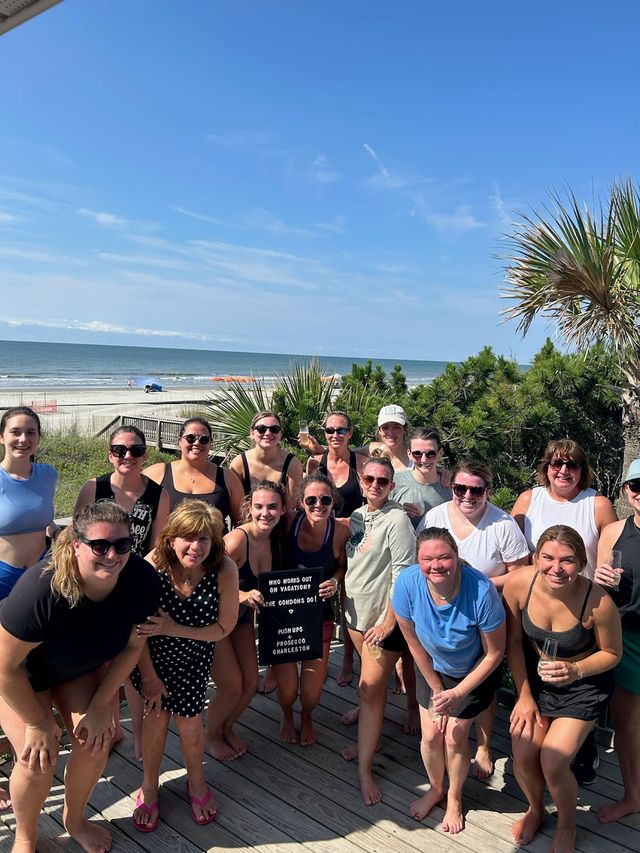 Smiling group of women on a sunny oceanfront wooden deck with palm trees and sandy beach in the background, one holding a small letter board — coastal Charleston beach vibe.