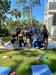 Group of women in activewear toasting and posing on yoga mats by a backyard pool, palm trees and a sunny coastal-style house in the background — outdoor fitness celebration.