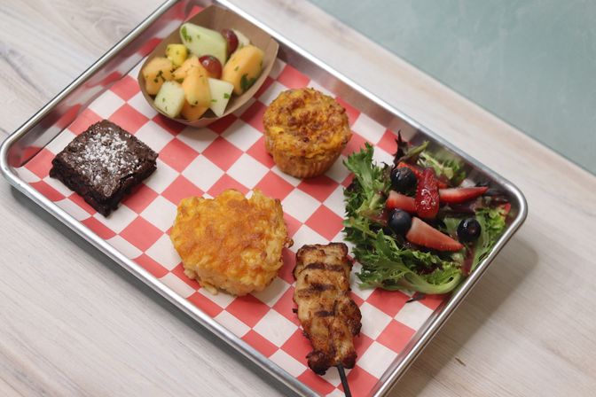 Metal serving tray with red-check paper holding a grilled chicken skewer, cheesy baked mac-and-cheese square, savory muffin, mixed green salad with strawberries and blueberries, a small fresh fruit cup, and a powdered-sugar brownie.