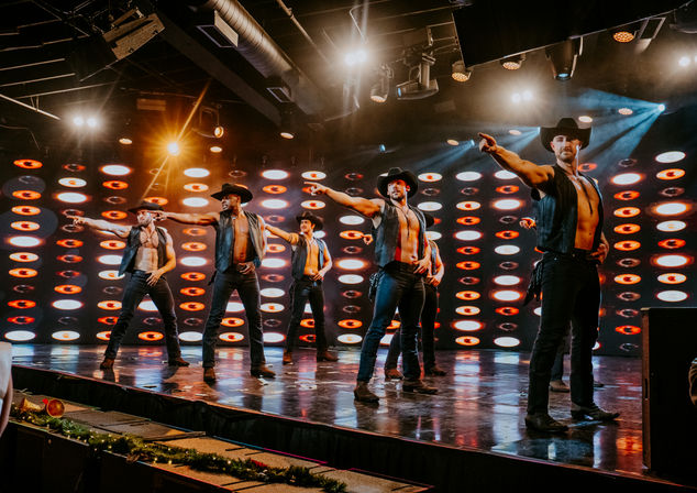 Shirtless male dancers in cowboy hats and leather vests performing an energetic choreographed routine on a nightclub stage with circular LED backdrop and dramatic spotlights.