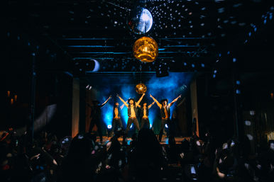 Five male dancers in open vests and hats strike a synchronized pose on a blue-lit nightclub stage beneath disco balls, with fog effects and a cheering audience in silhouette.