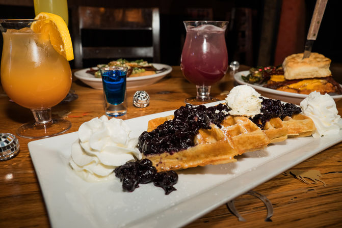 Waffle topped with blueberry compote, a scoop of vanilla ice cream and whipped cream on a white plate, set on a wooden table with colorful cocktails and a biscuit brunch plate in the background.