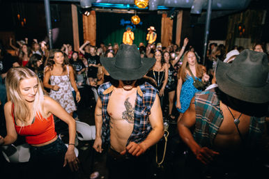 Rowdy country-nightclub scene with shirtless dancers in cowboy hats and plaid vests on a crowded dance floor, cheering crowd and performers on stage