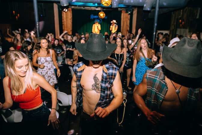 Rowdy country-nightclub scene with shirtless dancers in cowboy hats and plaid vests on a crowded dance floor, cheering crowd and performers on stage