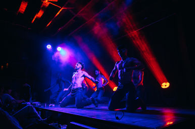 Shirtless male dancers kneeling on a nightclub stage performing an energetic choreography under dramatic red and blue spotlights and fog