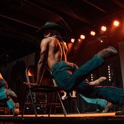 Shirtless dancer in a cowboy hat and jeans kicking off a folding chair during a high-energy stage performance under warm spotlights