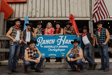 Smiling group of cowboy-clad performers and a woman posing outside a Nashville honky-tonk, wearing cowboy hats, flannel shirts and bandanas with an American flag visible
