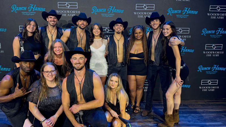 Group photo of country-themed performers and fans posing in front of a theater backdrop; men in cowboy hats and leather vests and women in dresses and boots smiling under stage lighting.