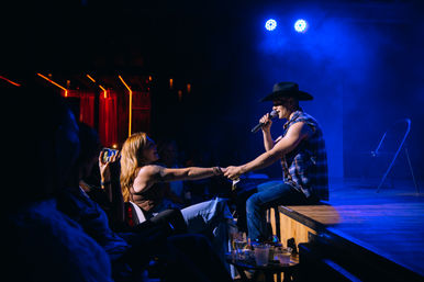Live country music performance in a dim nightclub: male singer in a cowboy hat and plaid shirt sits on stage under blue spotlights, holding a fan's hand while she records on her phone.