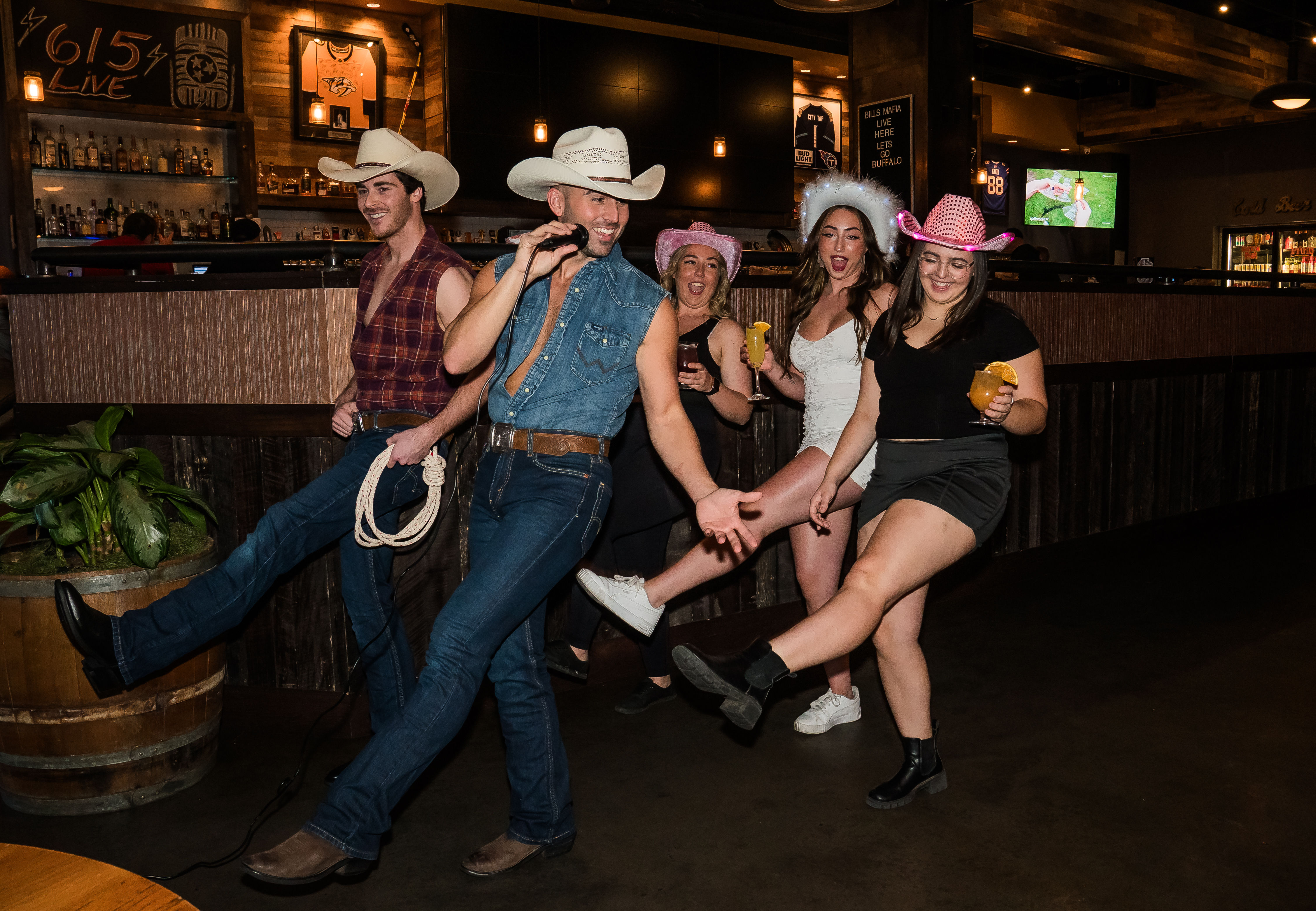 Group of five people in cowboy hats kicking their legs and singing together at a country-style bar, holding cocktails on a lively night out.