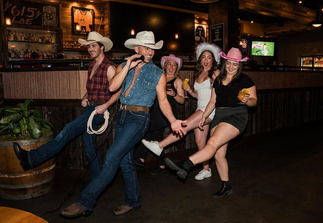 Group of five people in cowboy hats kicking their legs and singing together at a country-style bar, holding cocktails on a lively night out.