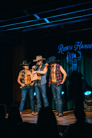 Three male country performers in cowboy hats, sleeveless flannel and denim on a lit wooden stage during a live country show, one holding a microphone and a box with audience silhouettes in the foreground.