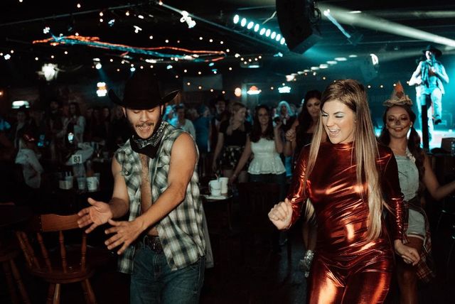 Crowded country-bar dance floor at night with a man in a cowboy hat and a woman in a metallic red outfit laughing and line-dancing under string lights and stage spotlights, live musician and cheering patrons in the background.