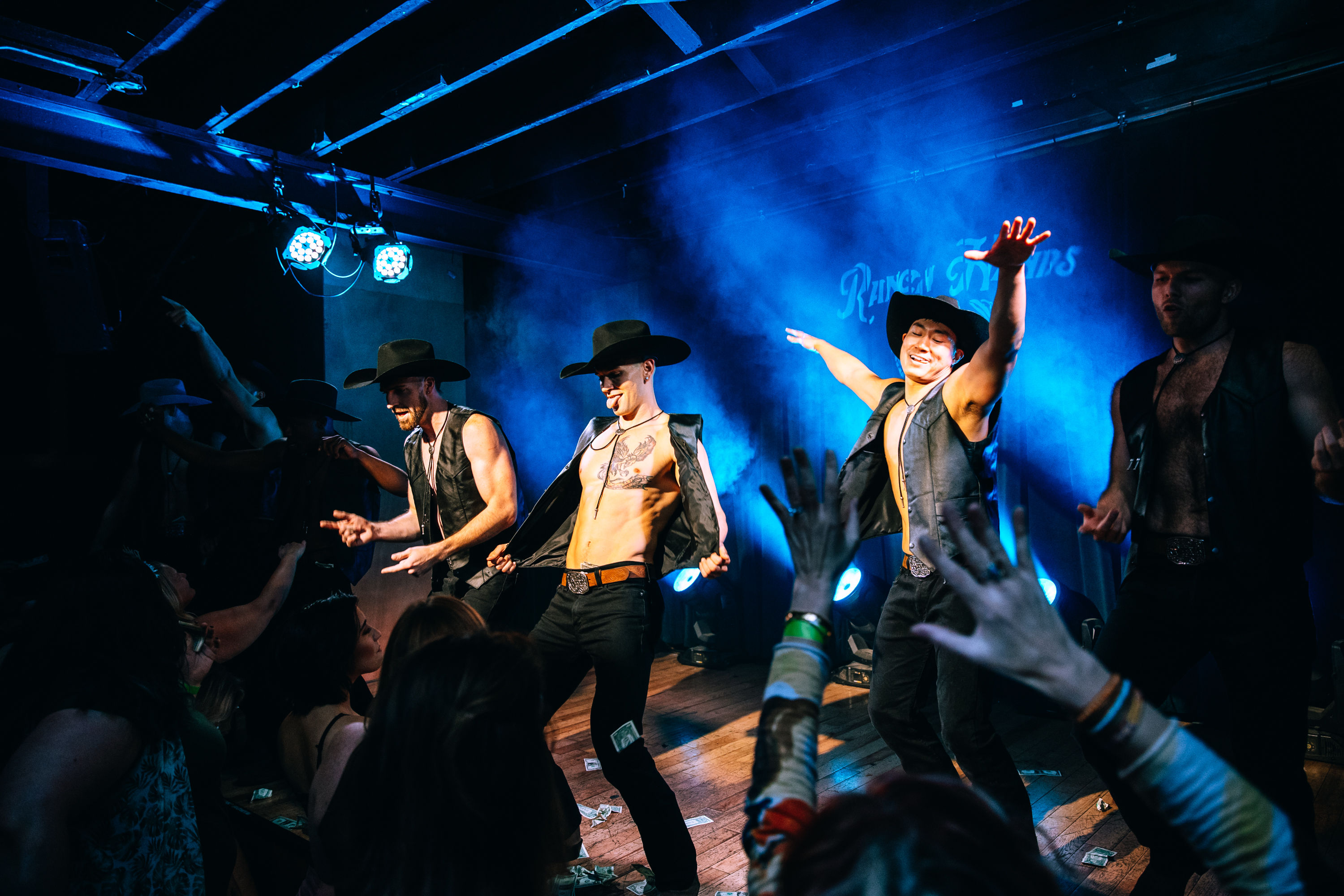 Energetic male cowboy dancers in open vests and hats performing a live male revue on a smoky blue-lit nightclub stage as an enthusiastic crowd reaches toward them, dollar bills scattered on the floor.