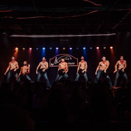 Shirtless male dancers in jeans and red bandanas pose with cowboy hats on a brightly lit Nashville stage, audience silhouettes in the foreground.