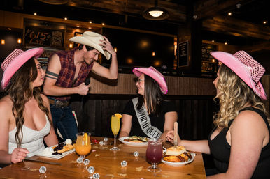 Three women in pink cowboy hats (one wearing a "Birthday Queen" sash) laugh as a cowboy-hatted singer with a microphone leans toward their wooden table of plates and cocktails in a rustic country-style bar.