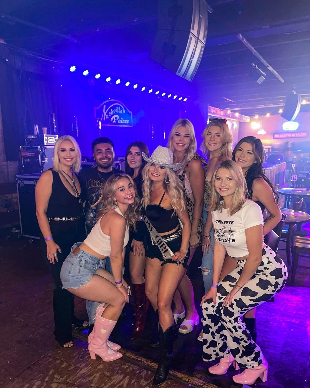 Smiling group of friends in cowgirl outfits and cowboy boots posing at a neon-lit Nashville country bar, one wearing a glitter cowboy hat and sash for a celebratory night out.