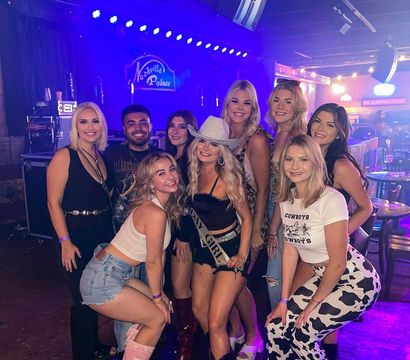 Smiling group of friends in cowgirl outfits and cowboy boots posing at a neon-lit Nashville country bar, one wearing a glitter cowboy hat and sash for a celebratory night out.
