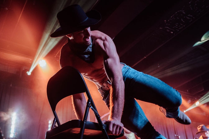 Shirtless male performer in a cowboy hat and bandana leans over a folding chair mid-dance on a smoky nightclub stage, striking a dynamic pose under warm red and blue spotlights.
