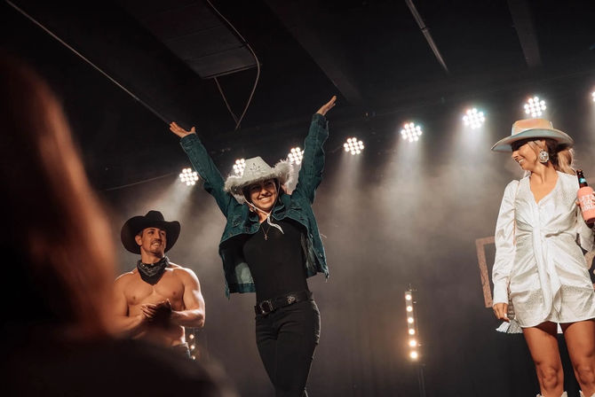 Live country music stage under spotlights with three performers: center person in a feathered white cowboy hat and denim jacket cheering with arms raised, shirtless cowboy clapping at left, woman in a white dress and cowboy hat holding a drink at right.