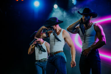 Three muscular male dancers wearing cowboy hats, bandanas, white tank tops and jeans performing a choreographed routine on a smoky stage with neon pink lighting.
