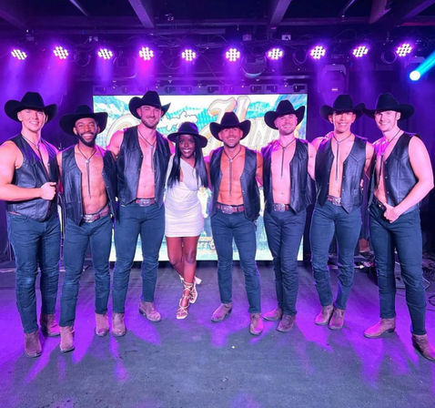 Group photo of eight shirtless male cowboy dancers in black hats, leather vests, jeans and boots flanking a woman in a white dress and strappy heels on a purple-lit stage with LED backdrop — nightclub cabaret entertainment.