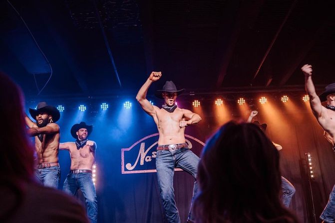 Shirtless male dancers in cowboy hats and jeans perform a country-themed stage routine under blue and orange lights with audience silhouettes in the foreground