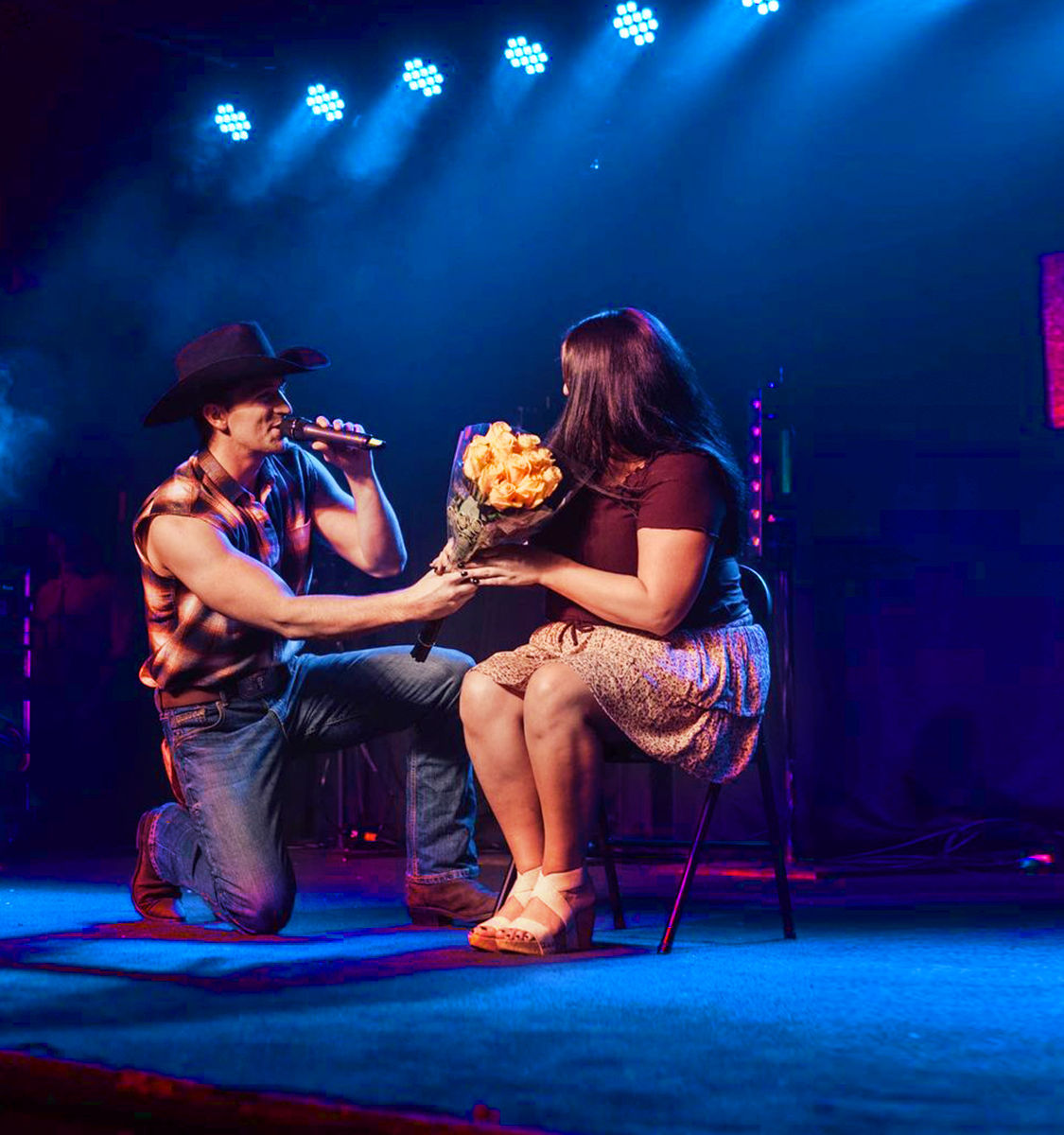 Cowboy-hatted singer kneeling on a blue-lit stage, offering a bouquet of yellow roses to a seated woman while singing into a microphone at a live country-music performance.