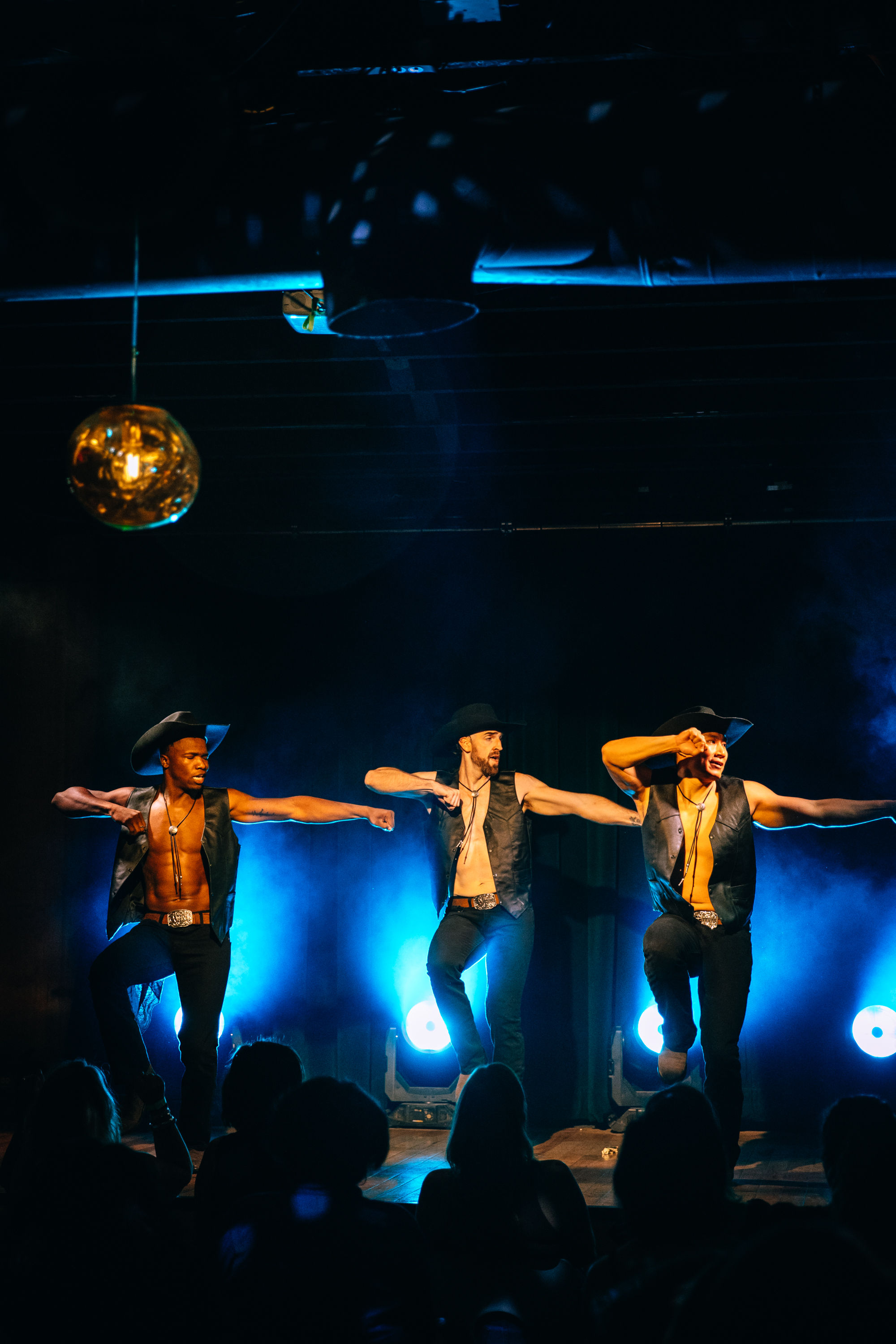 Three male dancers in cowboy hats and open vests mid-kick on a smoky nightclub stage with blue backlighting and a silhouetted audience watching.