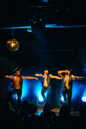 Three male dancers in cowboy hats and open vests mid-kick on a smoky nightclub stage with blue backlighting and a silhouetted audience watching.