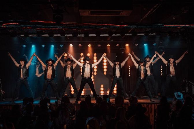 Shirtless male dancers in cowboy hats and leather vests strike a synchronized pose on a brightly lit nightclub stage with colorful spotlights and a silhouetted audience