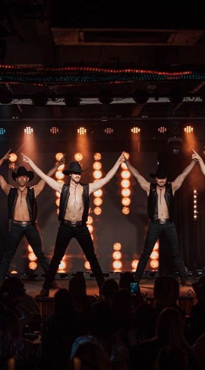 Shirtless male dancers in cowboy hats and leather vests strike a synchronized pose on a brightly lit nightclub stage with colorful spotlights and a silhouetted audience
