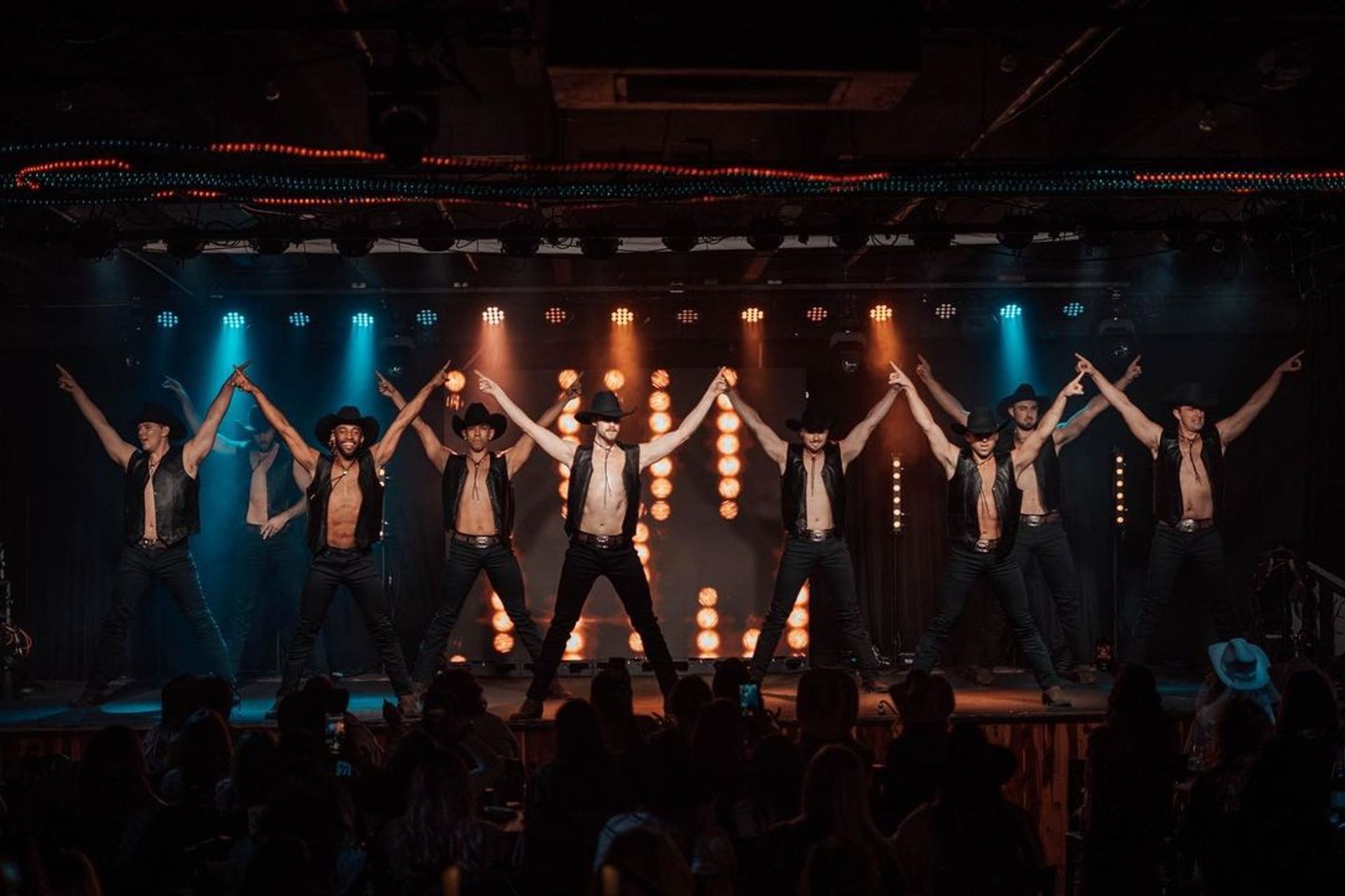 Shirtless male dancers in cowboy hats and leather vests strike a synchronized pose on a brightly lit nightclub stage with colorful spotlights and a silhouetted audience