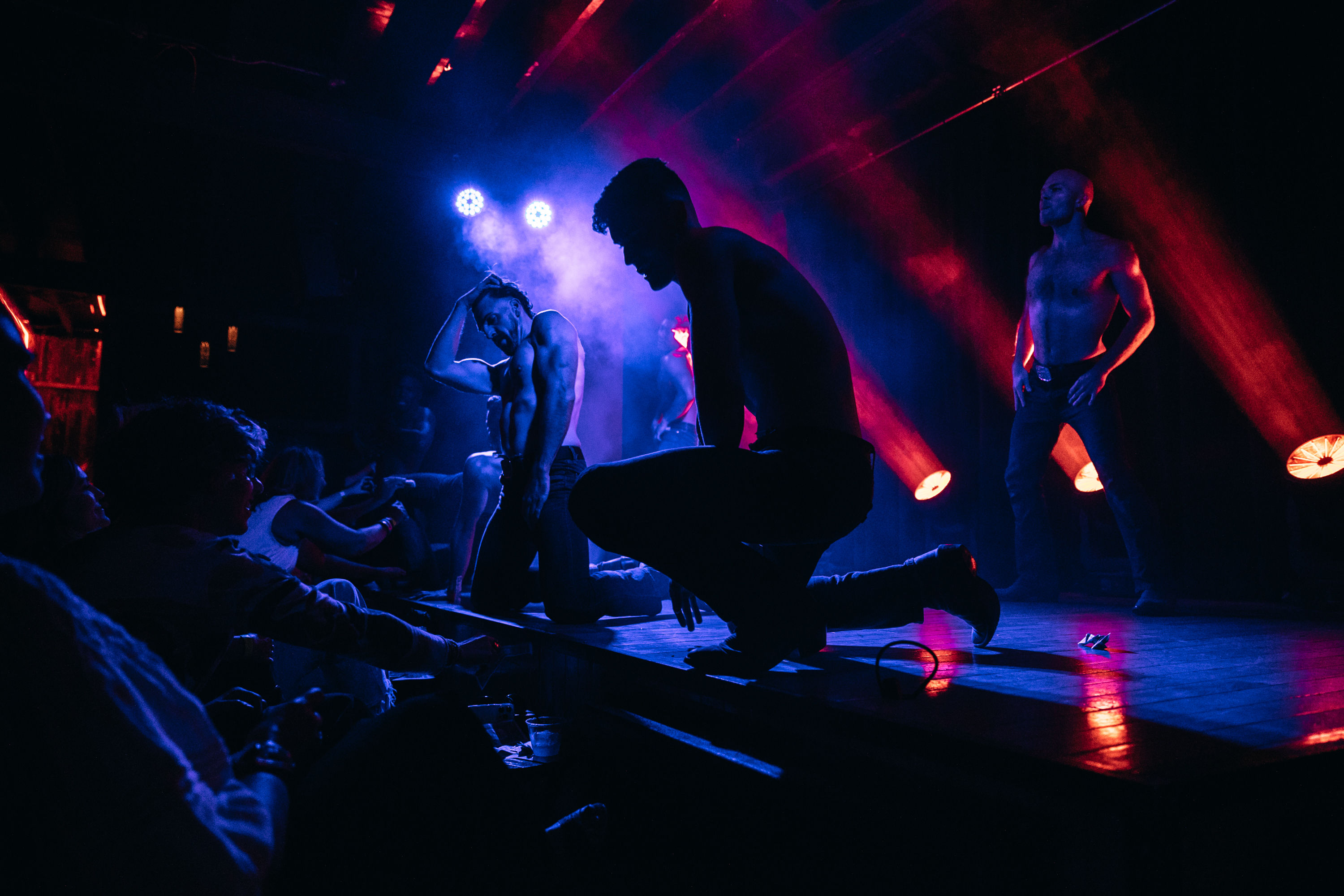 Shirtless male dancers performing on a dim nightclub stage with blue and red spotlights and fog, audience members reaching toward the stage