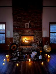 Cozy stone fireplace with roaring fire and twilight windows, arranged on a wooden floor with candles and sound‑healing instruments — gongs, crystal singing bowls, chimes and mallets.