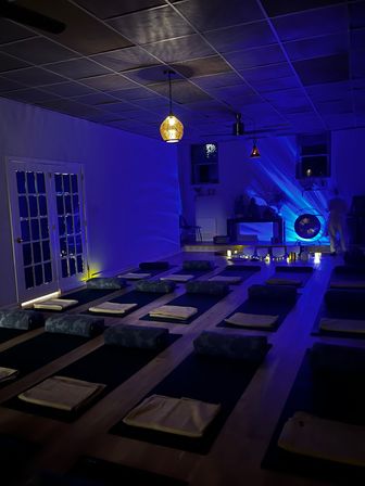 Blue-lit meditation studio with rows of floor mats, bolsters and folded blankets, a warm hanging pendant light, singing bowls and a gong at the front, candles creating an ambient sound-bath setup.