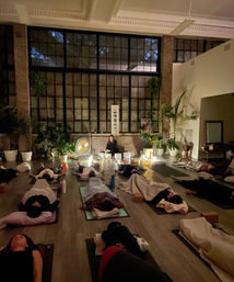 Evening sound bath in an urban loft yoga studio — students resting on mats under blankets while an instructor plays gong and crystal bowls by candlelight beneath large industrial windows and lush potted plants.