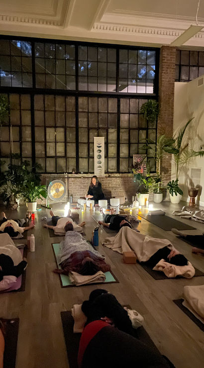 Evening sound bath in an urban loft yoga studio — students resting on mats under blankets while an instructor plays gong and crystal bowls by candlelight beneath large industrial windows and lush potted plants.