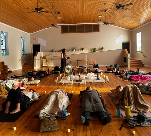 Calm group sound‑healing meditation in a sunlit wooden‑ceiling community hall — participants lying on mats and blankets around a central gong, crystal bowls, candles and crystals, with stained‑glass windows and hardwood floor.