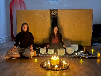 Two people seated cross-legged in a cozy, candlelit wellness studio surrounded by crystal singing bowls, mallets, fairy lights and a ring of candles on a wooden floor for a sound bath meditation.